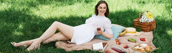 Mujer en vestido de verano sosteniendo melocotón cerca del vino y libros sobre hierba en el parque, pancarta - foto de stock