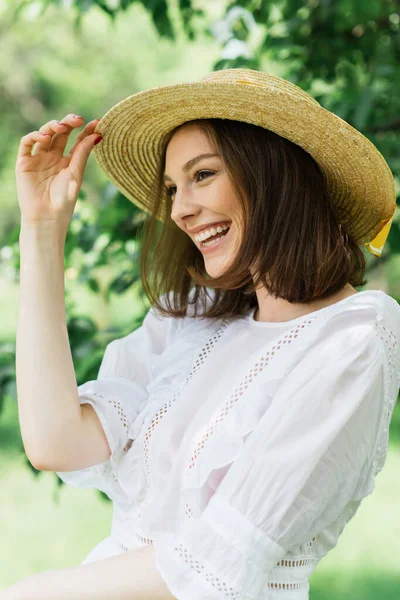 Mujer sonriente sosteniendo sombrero de paja y mirando hacia el parque - foto de stock