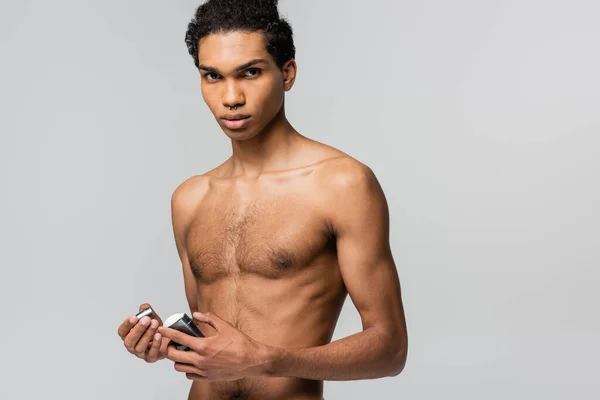 Joven afroamericano hombre mirando a la cámara mientras sostiene palo de antitranspirante aislado en gris - foto de stock