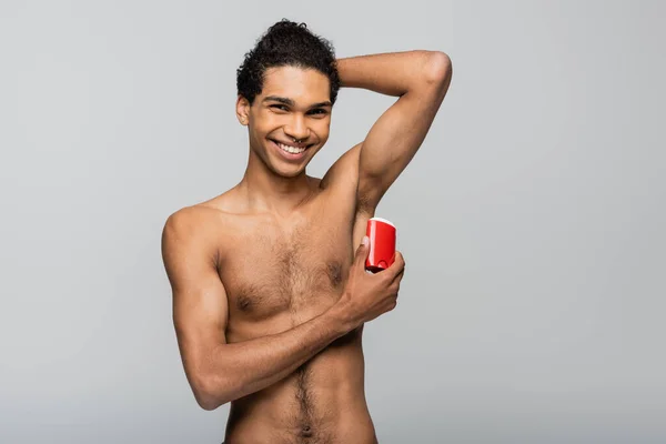 Happy african american man looking at camera while applying dry antiperspirant isolated on grey — Stock Photo