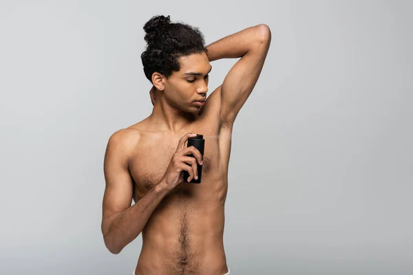 Sin camisa, joven afroamericano hombre rociando antitranspirante aislado en gris - foto de stock