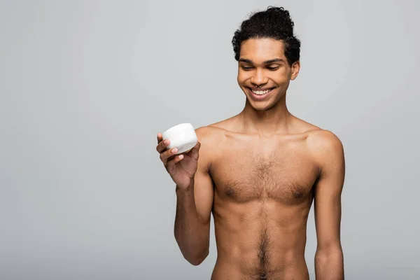 Веселий, без сорочки афроамериканський чоловік тримає контейнер косметичного крему ізольований на сірому — стокове фото