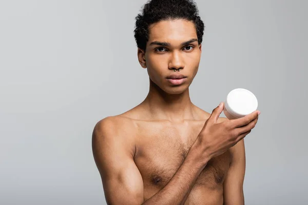 Африканский мужчина без рубашки показывает контейнер с косметическим кремом, изолированным на сером — стоковое фото