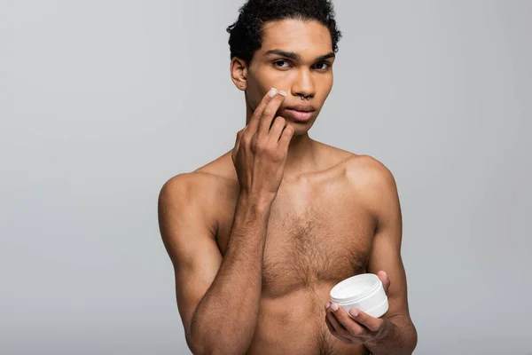 Молодий, без сорочки афроамериканський чоловік дивиться на камеру і наносить крем для обличчя ізольований на сірий — стокове фото