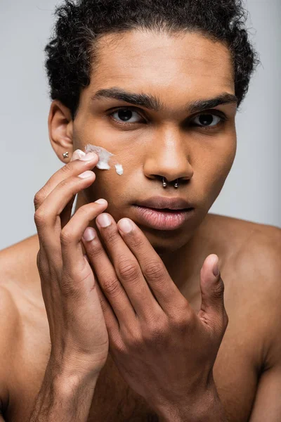Trafitto africano americano ragazzo applicando crema per il viso mentre guardando la fotocamera isolata su grigio — Foto stock