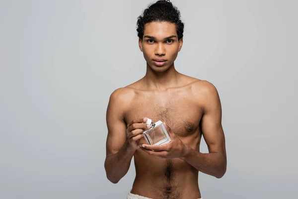 Joven, hombre afroamericano sin camisa mirando a la cámara mientras sostiene el perfume aislado en gris - foto de stock