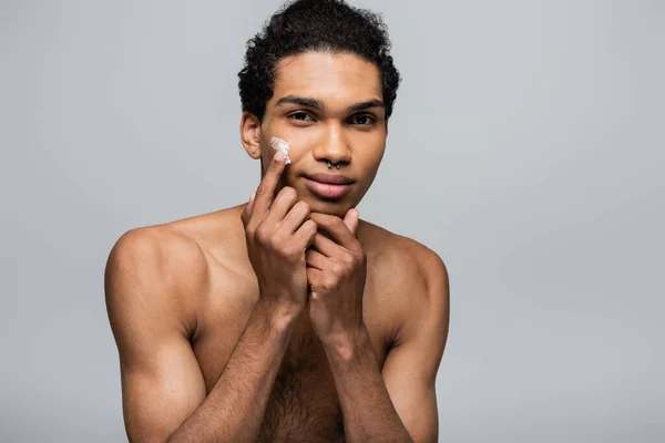 Joven afroamericano hombre aplicar crema facial mientras sonríe a la cámara aislado en gris - foto de stock
