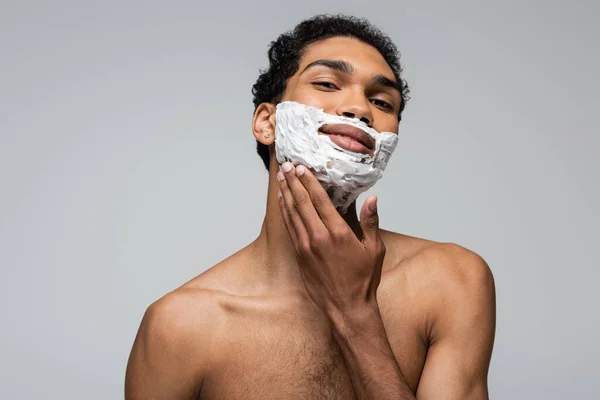 Africano americano chico mirando cámara mientras aplicación afeitado espuma aislado en gris - foto de stock