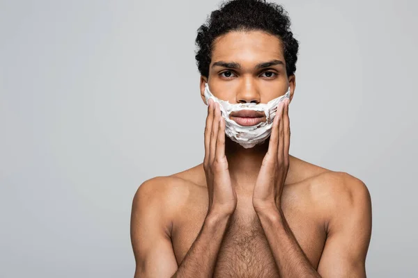 Vista frontal del hombre afroamericano aplicando espuma de afeitar en la cara aislada en gris - foto de stock