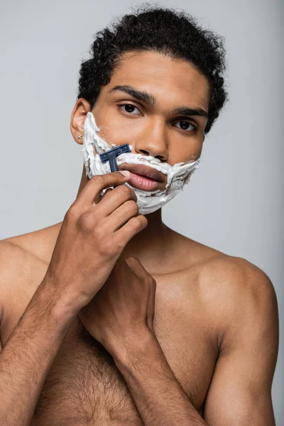 Hombre afroamericano mirando a la cámara mientras se afeita la cara con afeitadora de seguridad aislado en gris - foto de stock