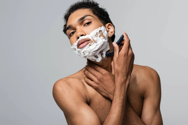 Giovane e senza maglietta uomo afroamericano guardando la fotocamera mentre la rasatura con rasoio di sicurezza isolato su grigio — Foto stock