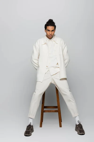 Full length view of african american man in white suit and sneakers posing near high stool on grey background — Stock Photo