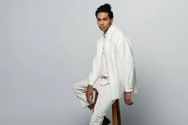 Joven afroamericano hombre en blanco elegante ropa sentado en taburete sobre gris fondo - foto de stock
