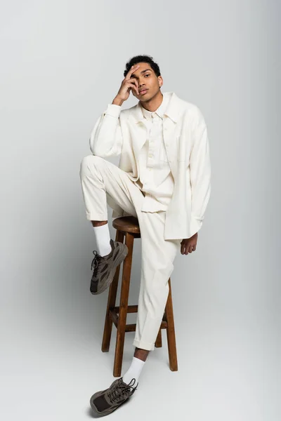 African american man in trendy clothes sitting on high stool with hand on face on grey background — Stock Photo