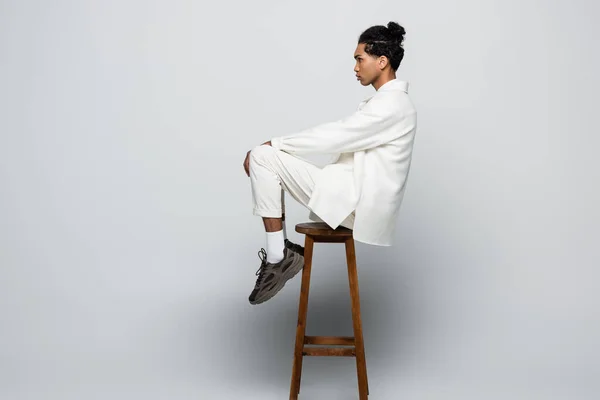 Vue latérale de l'homme afro-américain assis sur un tabouret haut en costume blanc et des baskets sur fond gris — Photo de stock