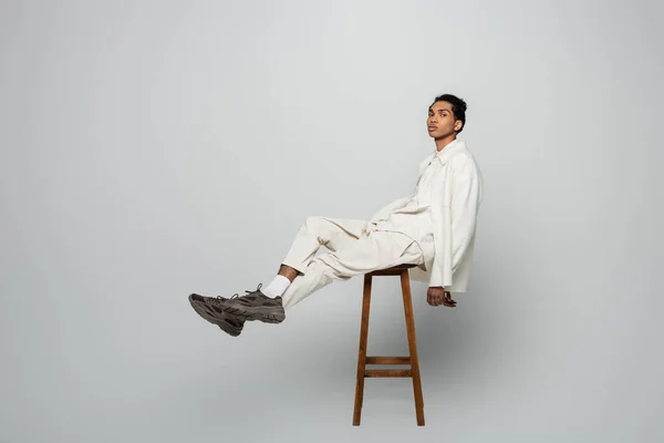 Africano americano hombre en blanco elegante ropa sentado en alto taburete en gris fondo - foto de stock