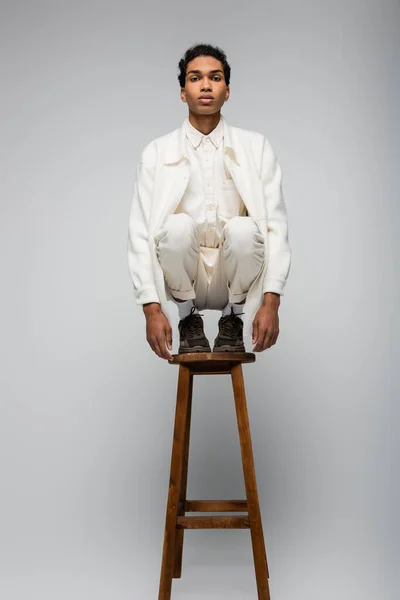 Front view of stylish african american guy in white suit and sneakers posing on high stool on grey background — Stock Photo