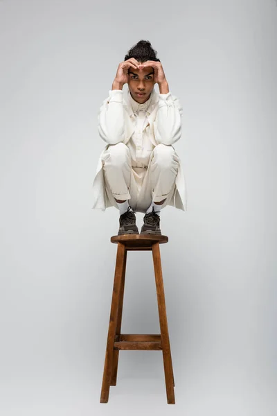 Stylish african american man in white suit and sneakers posing on high stool on grey — Stock Photo