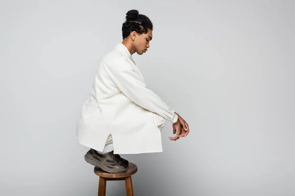 Side view of african american man on high stool in white jacket and sneakers on grey — Stock Photo