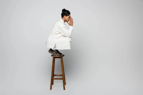 Vista laterale di ragazzo afroamericano in abiti bianchi in posa con le mani in preghiera su sgabello su sfondo grigio — Foto stock