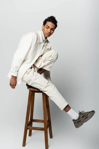 Joven afroamericano hombre en traje de moda y zapatillas de deporte posando en taburete alto sobre fondo gris - foto de stock