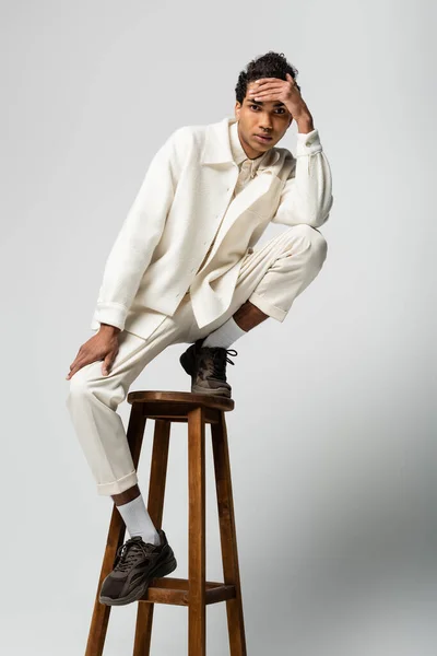 Afro-américain mec en vêtements blancs tenant la main près du front tout en posant sur un tabouret haut sur gris — Photo de stock