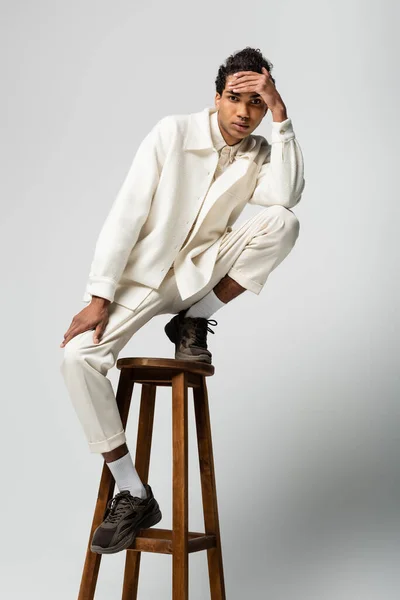 African american guy in white clothes holding hand near forehead while posing on high stool on grey — Stock Photo