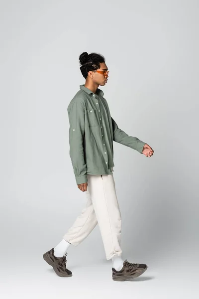 Vista laterale di elegante uomo africano americano in abiti casual e occhiali da vista a piedi su sfondo grigio — Foto stock