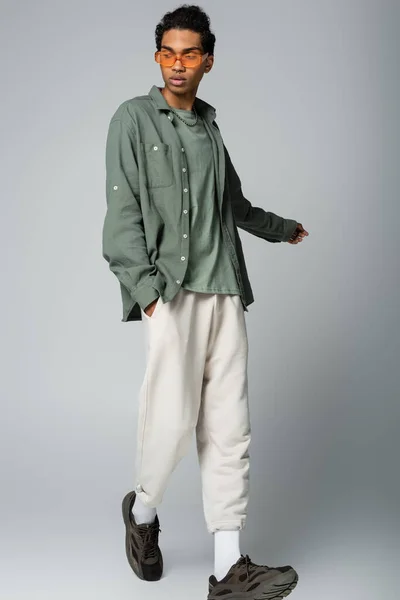 Stylish african american man walking with hand in pocket of white pants on grey background — Stock Photo