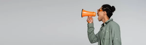 Seitenansicht eines trendigen afrikanisch-amerikanischen Mannes, der in einem Lautsprecher auf grau isoliert schreit, Banner — Stockfoto