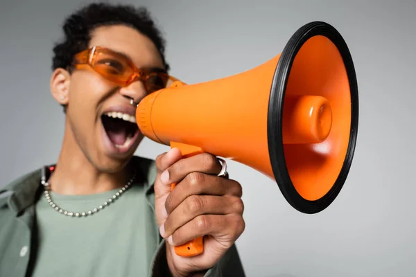 Blurred african american guy in orange eyeglasses screaming in loudspeaker isolated on grey — Stock Photo