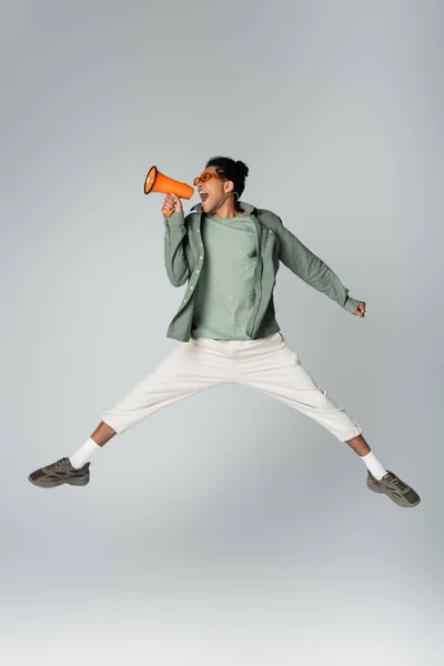 Homem americano africano em roupas da moda pulando com megafone em fundo cinza — Fotografia de Stock