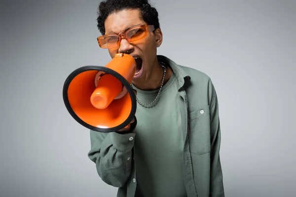 Giovane uomo afroamericano in occhiali alla moda urlando in megafono isolato su grigio — Foto stock