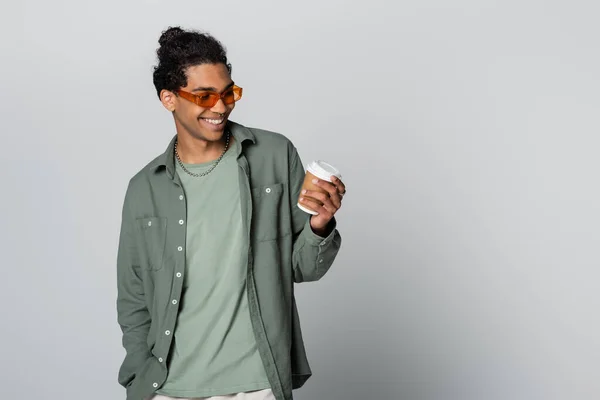 Joyful african american guy in stylish eyeglasses looking at paper cup isolated on grey — Stock Photo