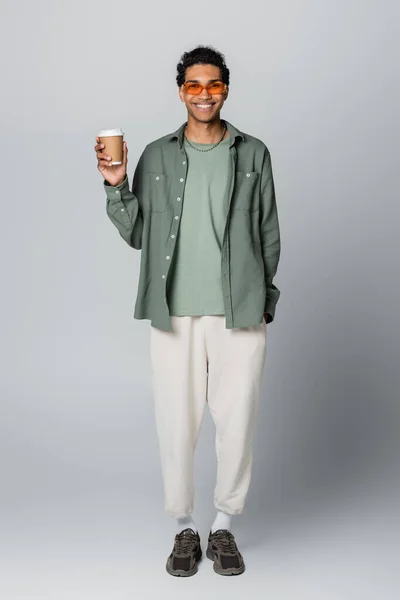 Full length view of african american guy in trendy clothes holding takeaway drink isolated on grey — Stock Photo