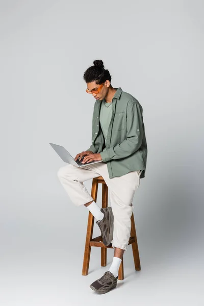 Homem americano africano na moda digitando no laptop enquanto sentado em alto banco em fundo cinza — Fotografia de Stock