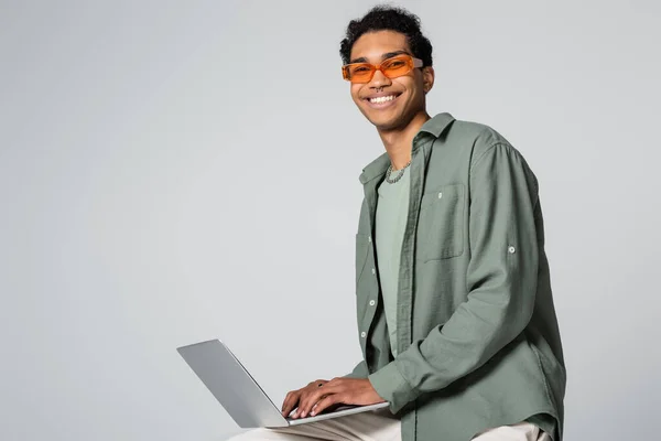 Cheerful african american guy in stylish eyeglasses typing on laptop and smiling at camera on grey background — Stock Photo