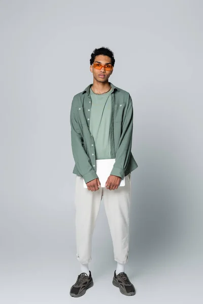 Vista integrale dell'uomo afroamericano alla moda in piedi con laptop su sfondo grigio — Foto stock