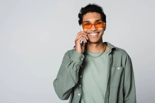 Alegre afroamericano hombre sonriendo a la cámara mientras habla en el teléfono celular aislado en gris - foto de stock