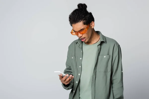 Chocado afro-americano homem em óculos laranja olhando para celular isolado em cinza — Fotografia de Stock