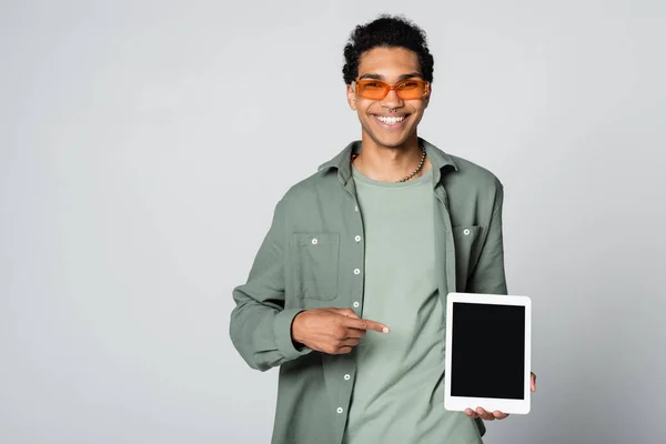 Веселый африканский американец в оранжевых очках, указывающий на цифровой планшет, изолированный на сером — стоковое фото