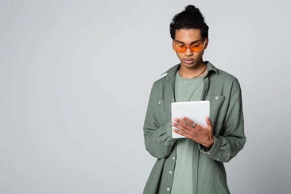 Молодой африканский американец в стильной рубашке и очках, используя цифровой планшет, изолированный на сером — стоковое фото