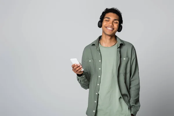 Ragazzo afroamericano con smartphone ascoltare musica mentre tiene la mano in tasca isolato su grigio — Foto stock