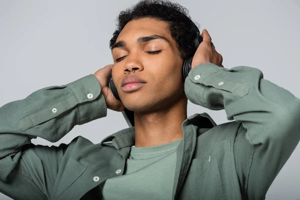 Junger afrikanisch-amerikanischer Mann berührt Kopfhörer, während er mit geschlossenen Augen Musik hört, isoliert auf grau — Stockfoto