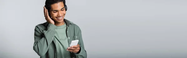 Joyeux homme afro-américain regardant le téléphone portable tout en écoutant de la musique isolée sur gris, bannière — Photo de stock