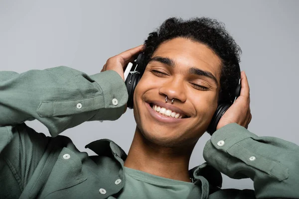 Feliz afro-americano cara em fones de ouvido ouvir música com olhos fechados isolado em cinza — Fotografia de Stock