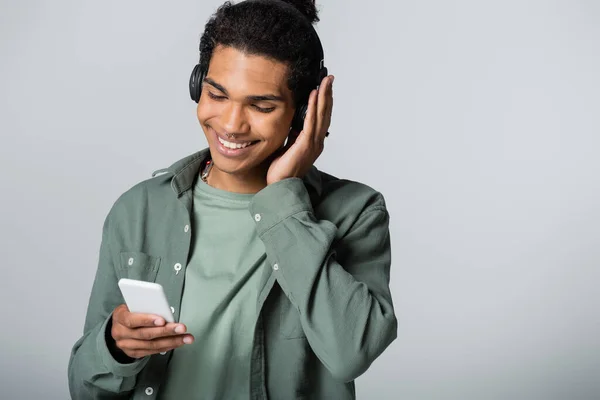 Stylish african american guy looking at smartphone while listening music in wireless headphones isolated on grey — Stock Photo