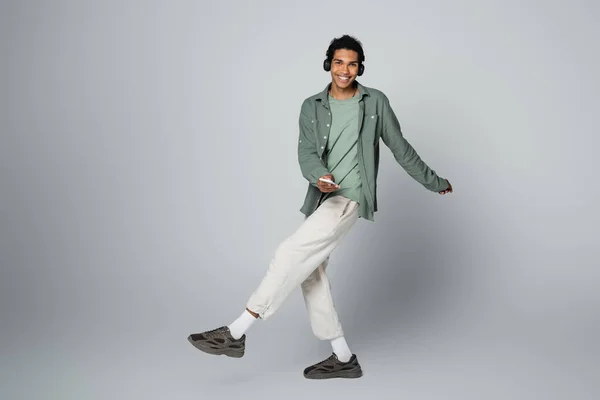 Hombre afroamericano feliz en ropa elegante escuchando música y bailando aislado en gris — Stock Photo