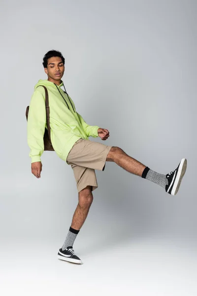 Homem americano africano na moda com capuz e shorts posando em uma perna isolada no cinza — Fotografia de Stock