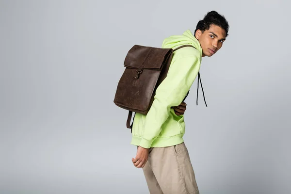 Hombre afroamericano de moda con marrón, mochila de cuero mirando a la cámara aislada en gris - foto de stock